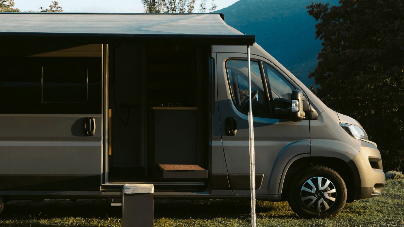 Furgoneta camper gris con el toldo desplegado sobre la hierba, en un prado de montaña al atardecer.